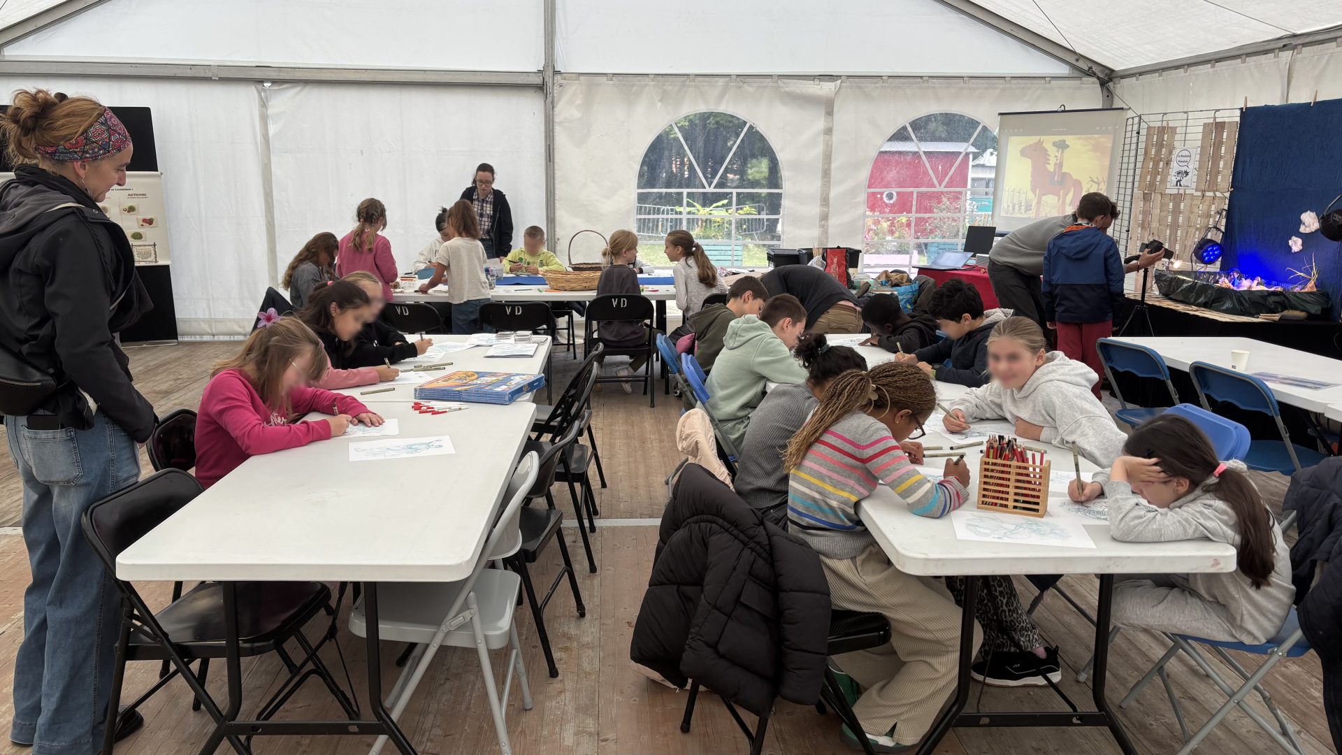 Groupe d’enfants participant à un atelier de dessin dans une grande tente, assis à des tables avec du matériel de création, accompagné·es d’adultes encadrants.